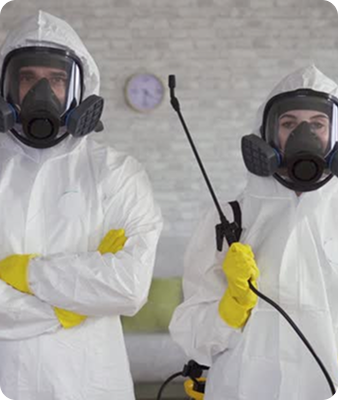Deux personnes portant des combinaisons blanches, des gants jaunes et des masques respiratoires se tiennent à l'intérieur. L'une d'elles tient une lance de pulvérisation, ce qui suggère un travail de désinfection ou de décontamination. Une horloge est accrochée au mur derrière eux.