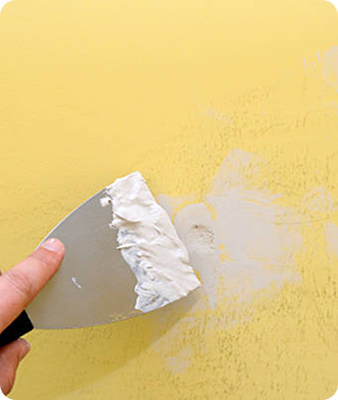 Une main utilise un couteau à mastic pour étaler du mastic blanc sur un petit trou dans un mur jaune, réparant ainsi la surface.