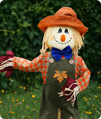 Un joyeux épouvantail au visage souriant, avec un chapeau orange, un nœud papillon bleu, une chemise à carreaux et une salopette verte ornée d'une feuille de tissu, se tient dans une zone herbeuse sur fond vert et feuillu.