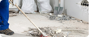 Une personne en pantalon bleu balaie des débris de construction sur un sol poussiéreux. À l'arrière-plan, des piles de gravats, des sacs poubelles, une pelle et des câbles apparents sur un mur indiquent que des travaux de rénovation sont en cours.