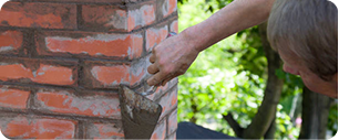 repa Une personne utilise une truelle pour appliquer du mortier entre les briques d'un mur de briques rouges, avec un feuillage vert visible à l'arrière-plan.
