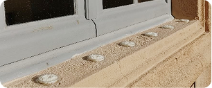 Un rebord de fenêtre beige avec des pics anti-pigeons ronds, blancs et texturés, espacés régulièrement sur sa surface, placé sous un cadre de fenêtre gris.