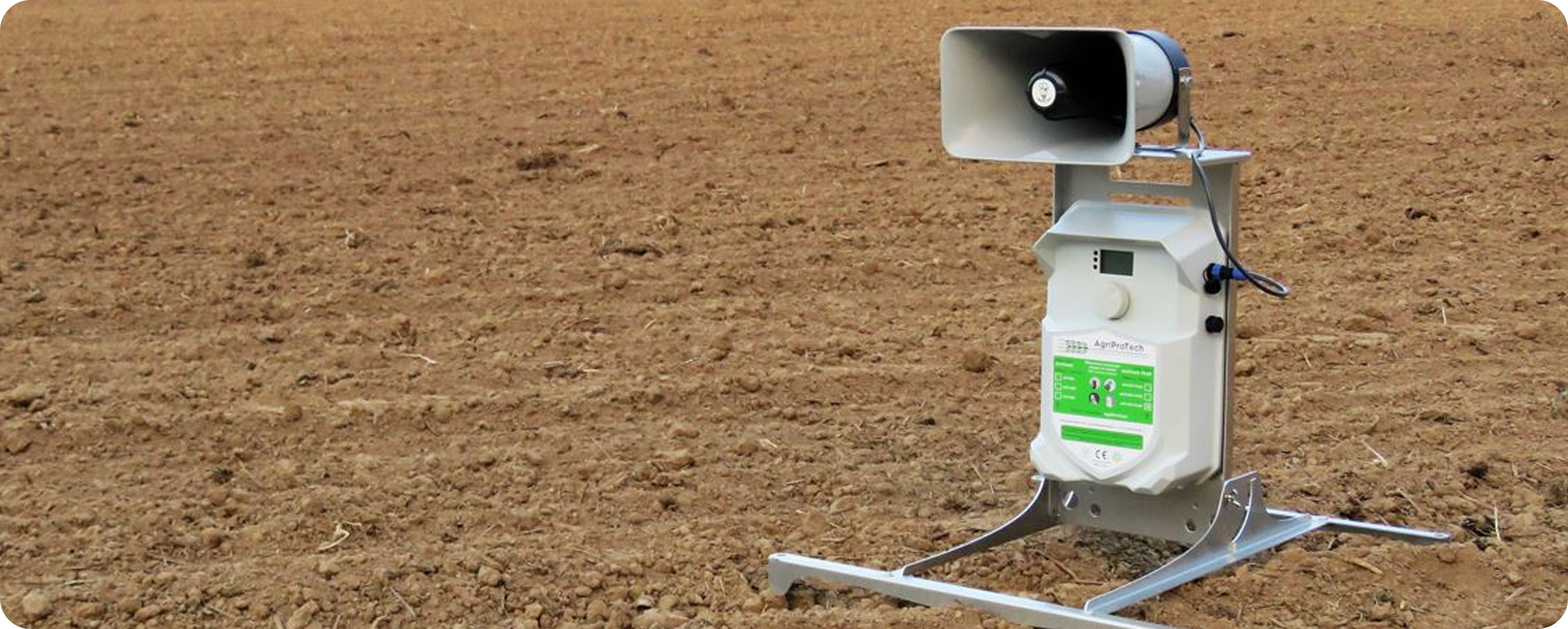 Un dispositif électronique alimenté par l'énergie solaire, doté d'un haut-parleur et d'un panneau de commande, est posé sur un cadre métallique dans un champ agricole labouré. Il est probablement utilisé pour dissuader les oiseaux ou les animaux.