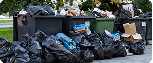 De grandes piles de sacs poubelles et de déchets en vrac débordent des bennes à ordures sur le sol, tandis que des cartons et divers déchets sont éparpillés dans un espace extérieur.